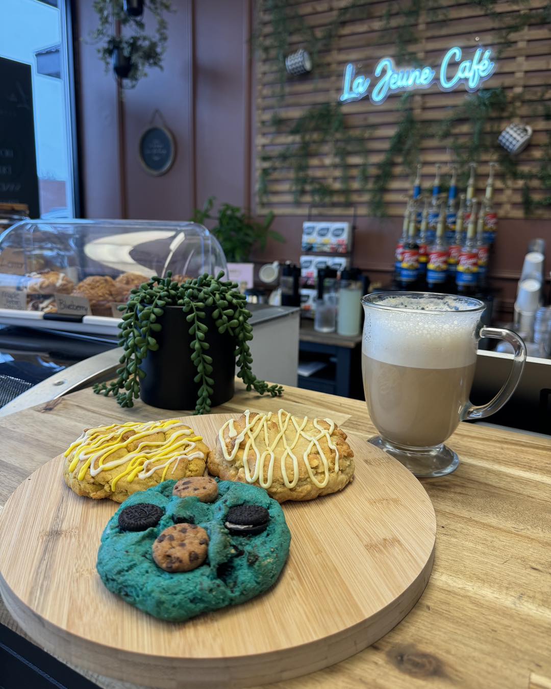 a wooden tray of cookies sits on a counter in front of a latte and a vine-covered neon sign that reads "La Jeune Café".
