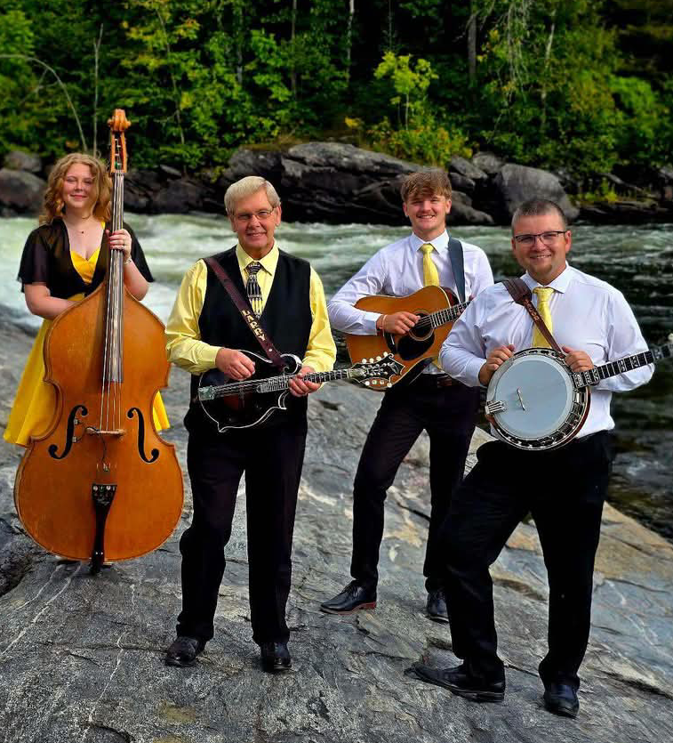 Larry Efaw and the Bluegrass Mountaineers, a smiling bluegrass band neatly dressed in yellow, white and black dress clothes, posing with their instruments on a rocky river bank in front of a lush green forest.