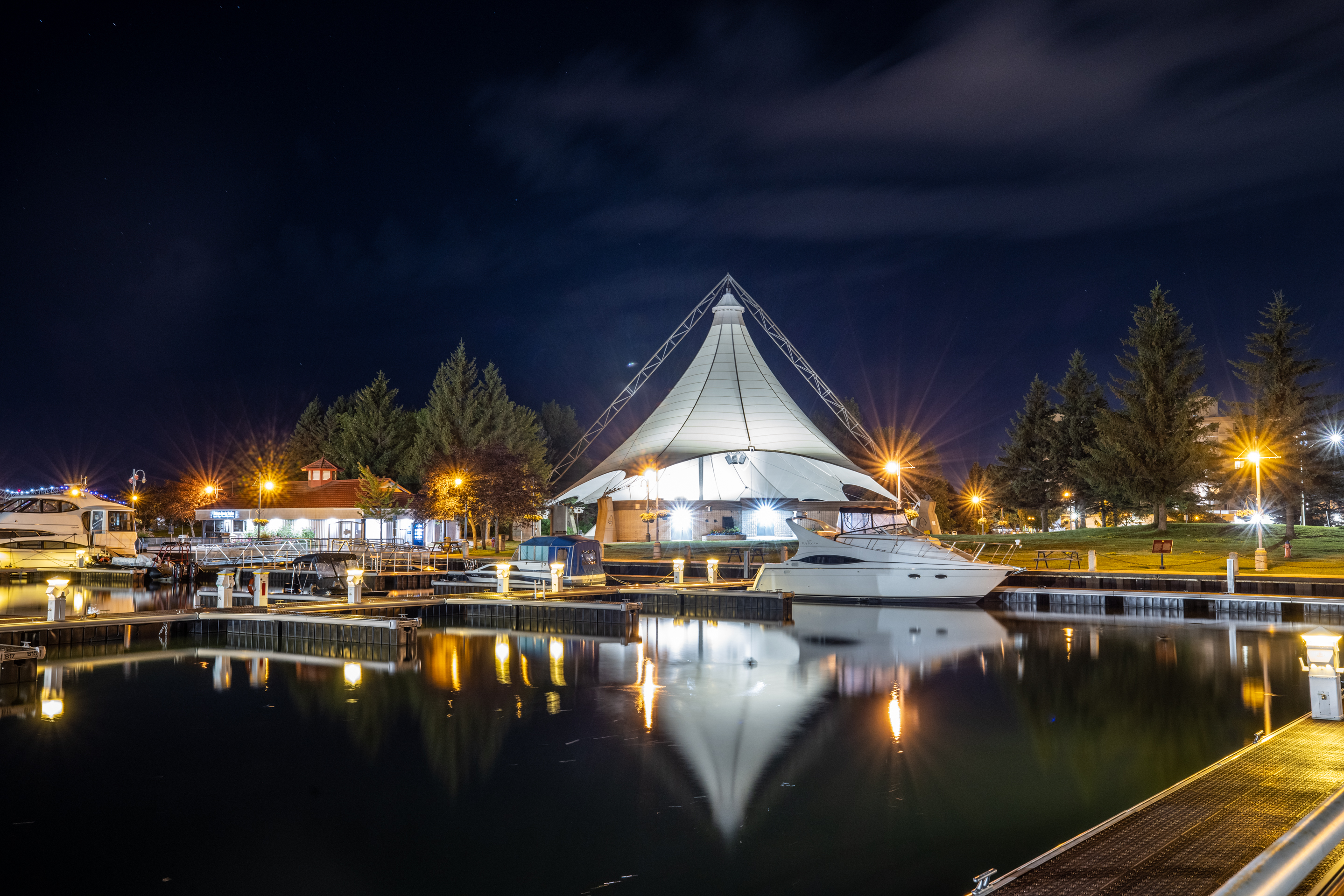 The white spire of the Roberta Bondar Pavilion at night, illuminated from within and surrounded by sparkling city lights reflecting mirror-like in the dark lake water next to it. 