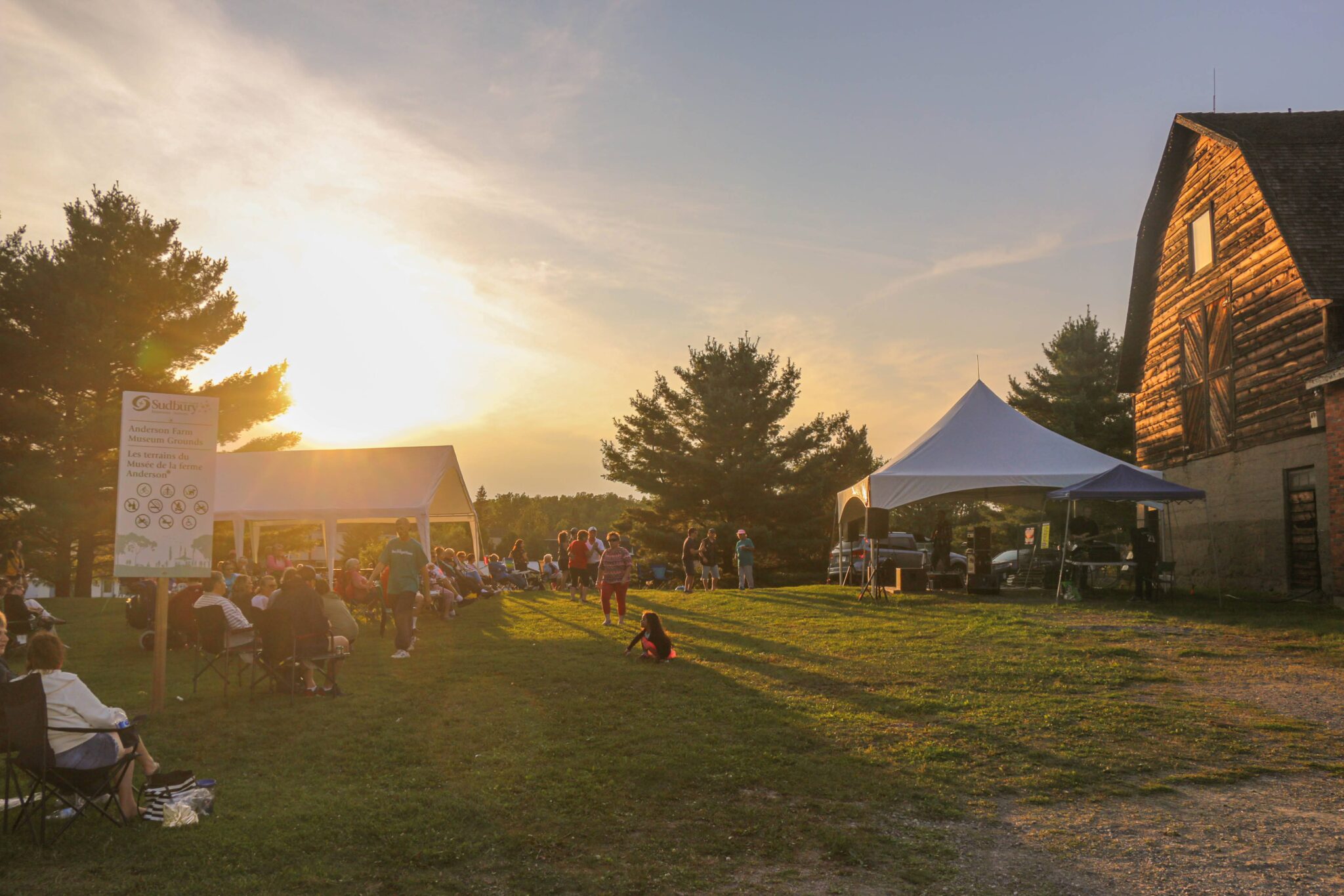 The sun sets behind a green forest and field and a series of market tents around a large old wooden barn at the Rock the Farm event. 