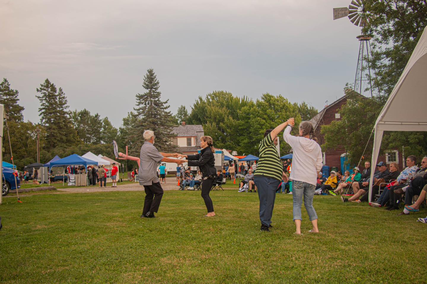 People dance happily together in a grassy clearing surrounded by canvas market tents, green forest and an aged wooden barn on a golden summer evening at Rock the Farm.