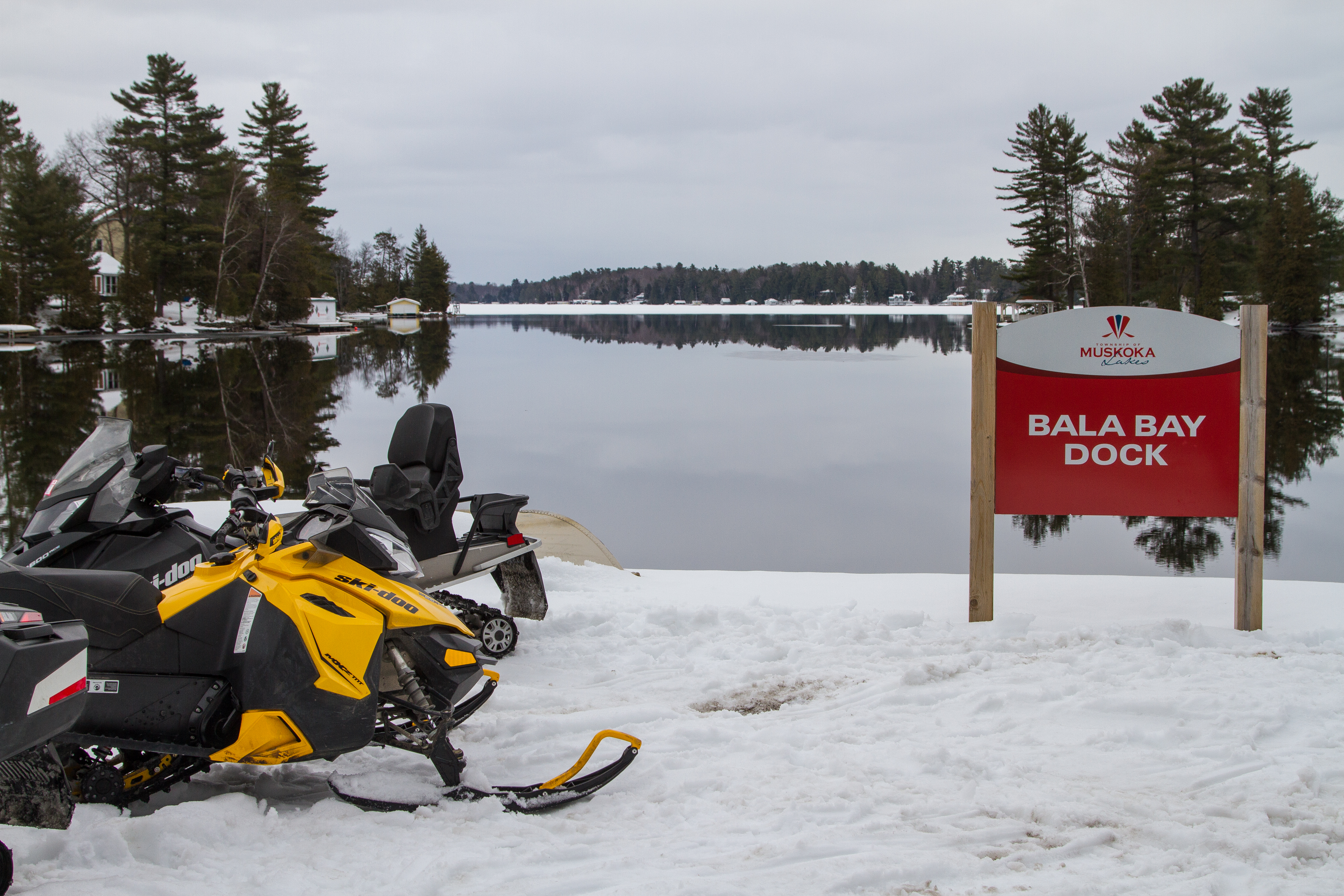 a sign next to an open lake surrounded by snowy banks reads "Bala Bay Dock, Muskoka Lakes". A couple of snowmobiles sit parked on the shore next to it.