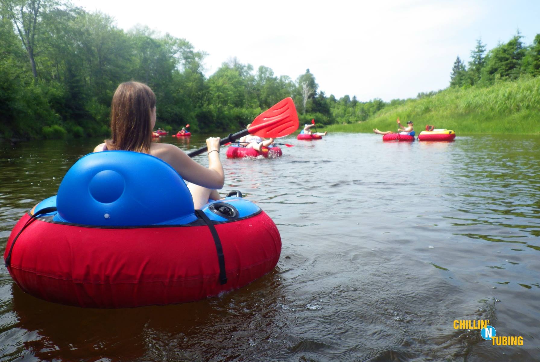Several people in red inner tubes float along a lazily moving river surrounded by green forest on a summer day. 
