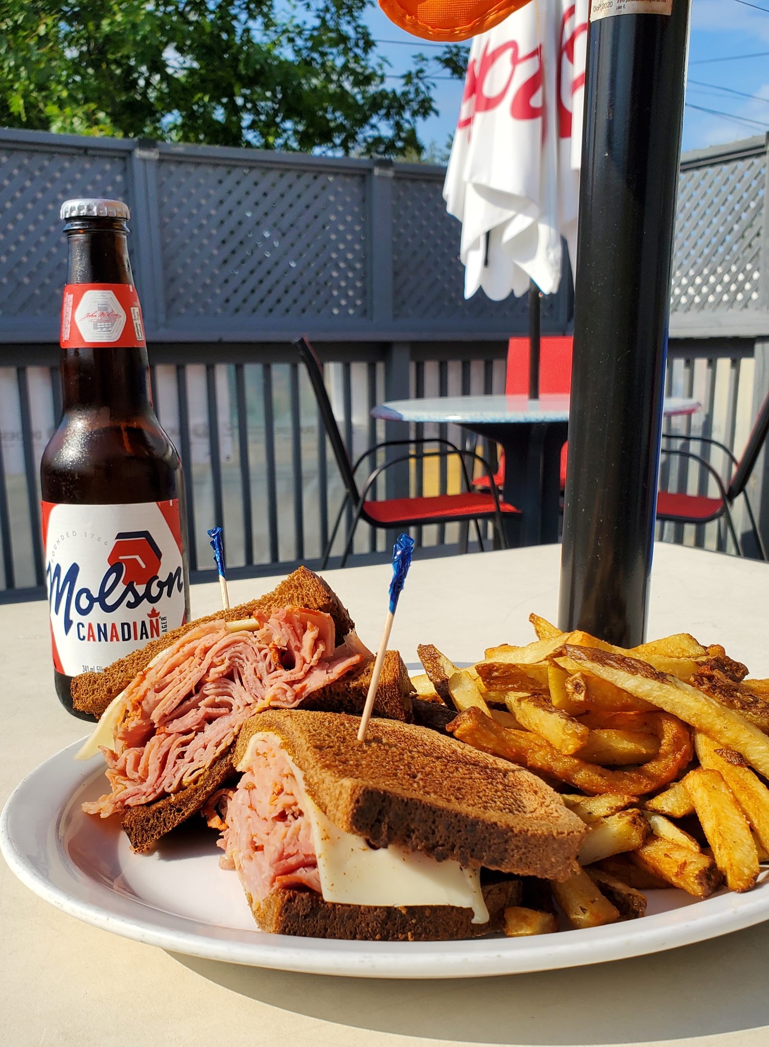 A reuben sandwich and fries with a bottle of beer on a patio table under blue sky and a green tree at the Doghouse Azilda.