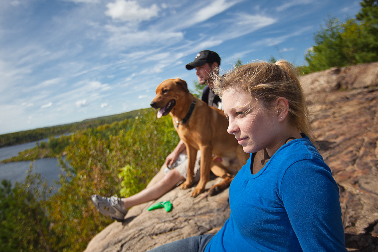 a man and woman sit with their dog on a rock lookout, gazing out contentedly over a high valley view on a breezy summer day in Sudbury.