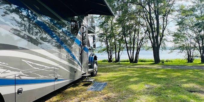 an RV parked under a shady canopy of green trees on a lawn of soft spring grass looking out over a calm blue lake.