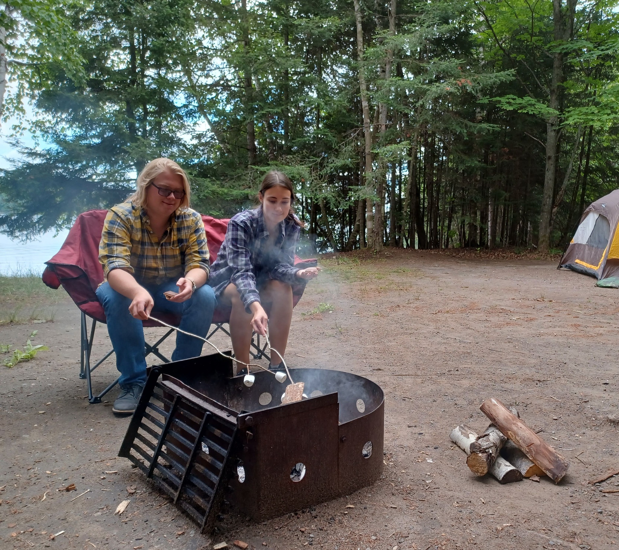 two people smile as they toast marshmallows next to a campfire at a tidy, treed campsite at Fairbank Provincial Park. 