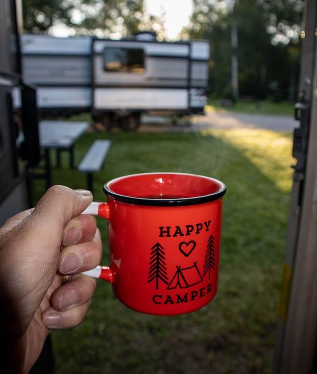 A hand holds a red mug with a "Happy Camper" print on it in front of the open door of an RV. Lush green grass, an RV campground and a rising sun are outside.