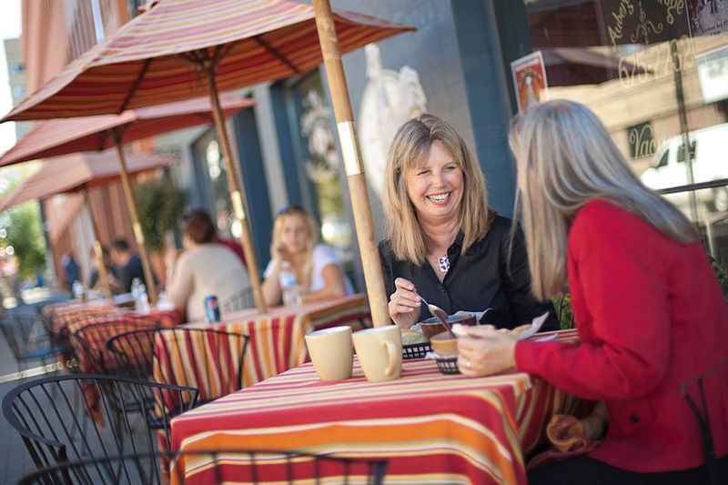 two older women smile and chat over coffee at a covered patio table on a summer day. 