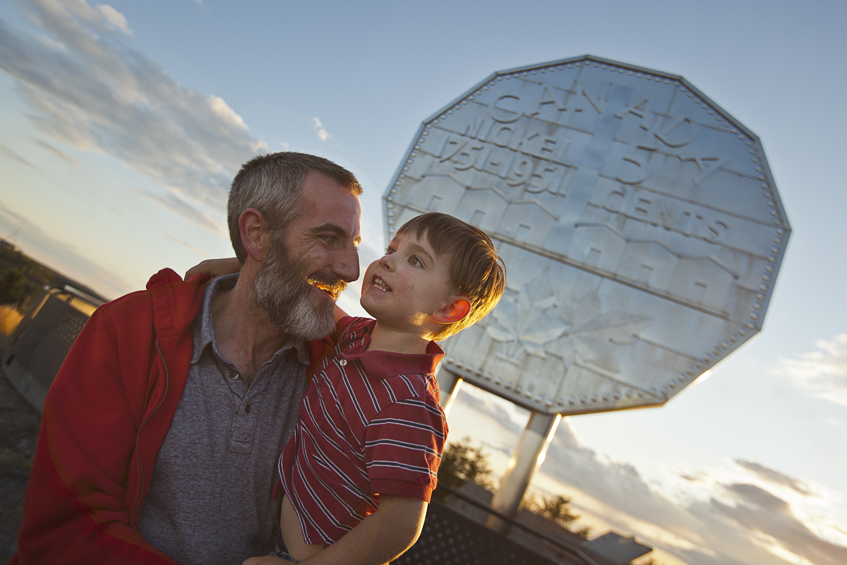 an older man smiles at the young boy that he is holding as they stand together in front of the Big Nickel at sunset in Sudbury. 