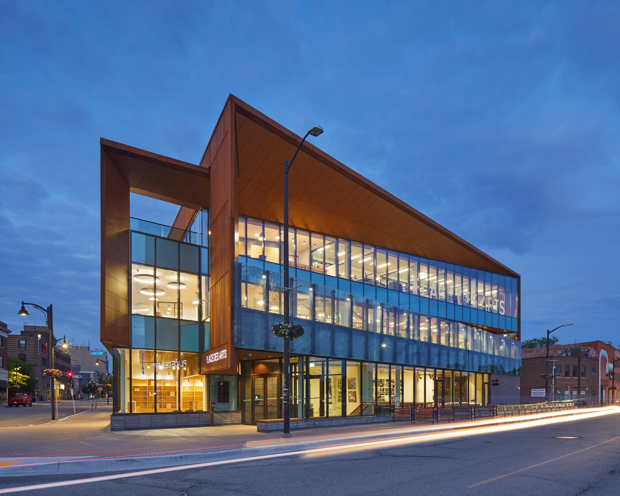 Place des Arts in Sudbury, a sleek, glass-fronted building lit up at twilight. 