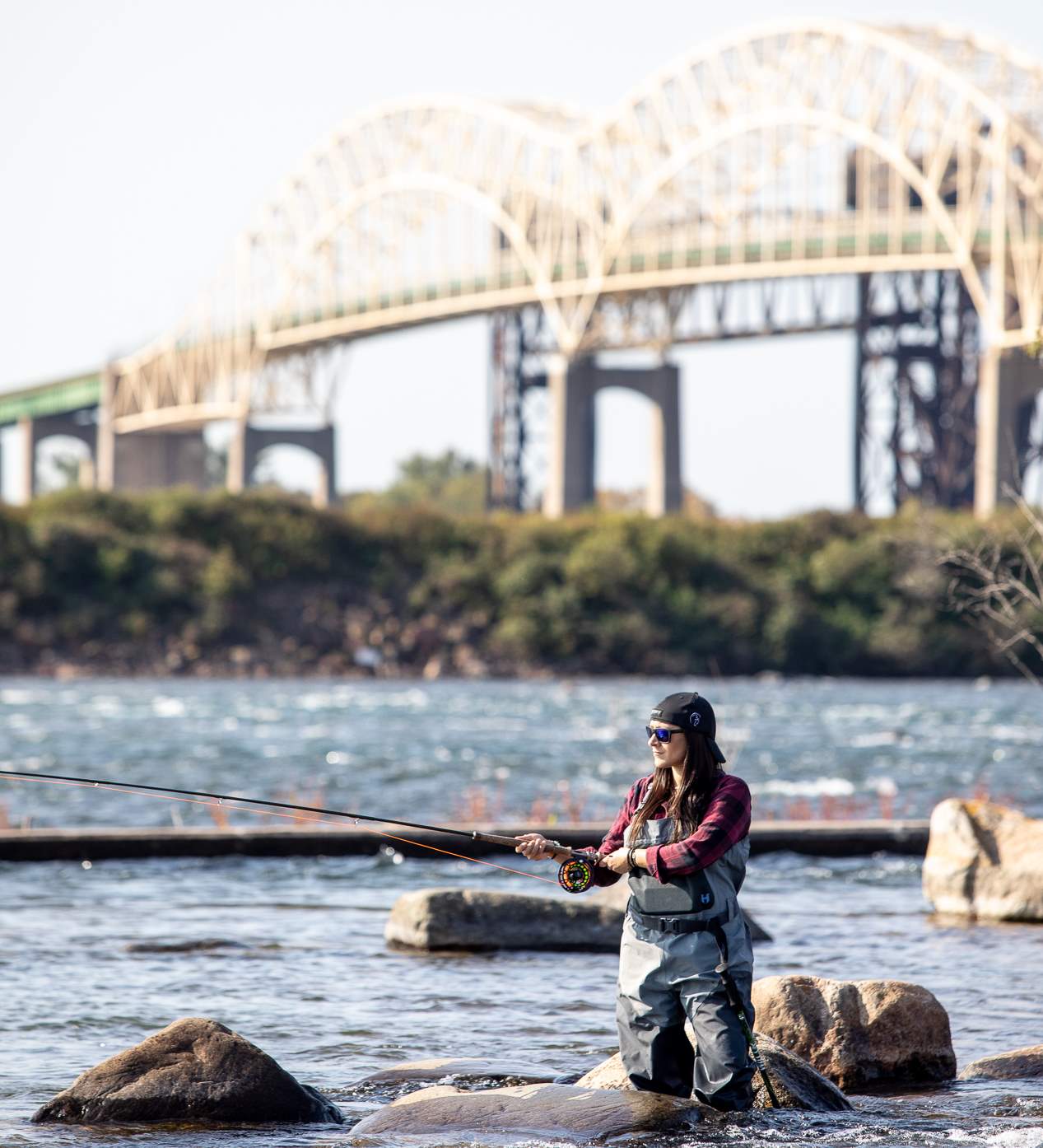 an angler casts into the shining waters of the wide St. Marys River on a sunny summer day. The International Bridge is in the background.