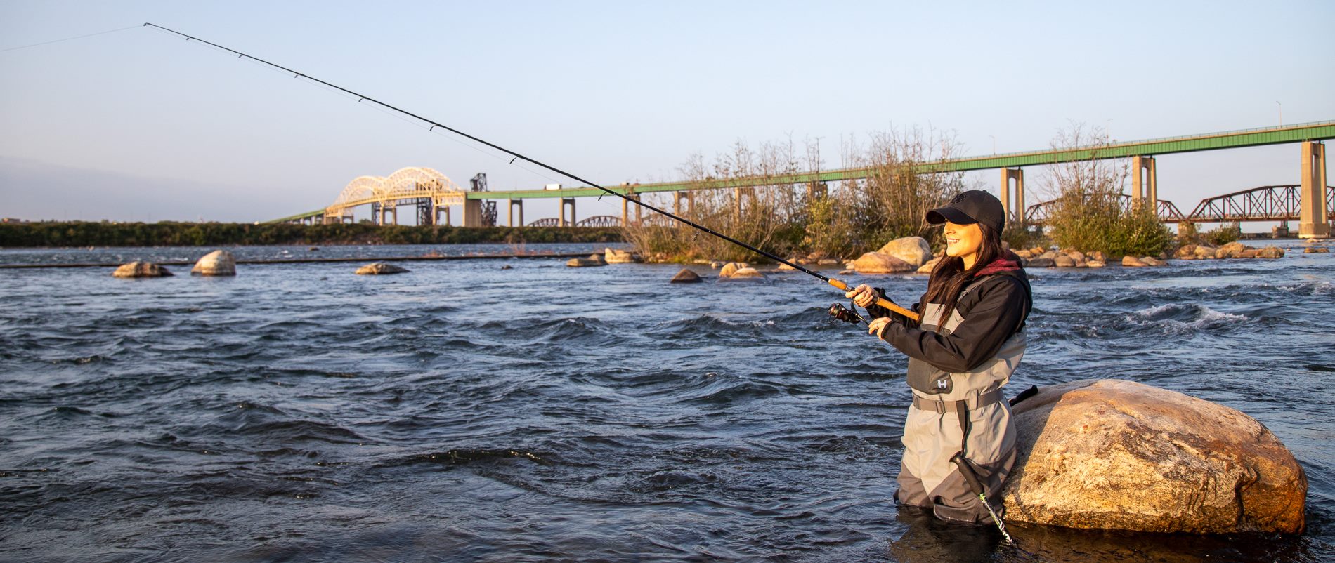an angler smiles as they reel, standing knee-deep in the water of the St. Marys Rapids on a sunny spring day. The International Bridge is in the background. 