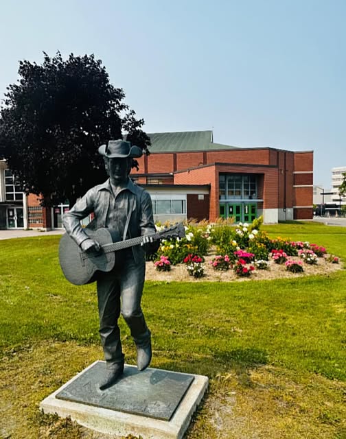 a statue of the musician Stompin' Tom Connors playing the guitar and wearing a cowboy hat surrounded by green grass and flowers.