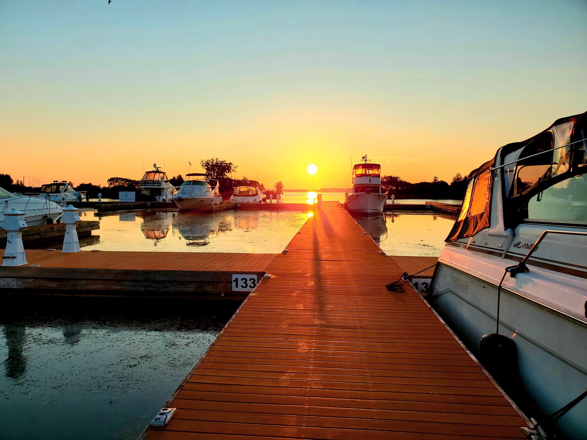 boats sit in the slips on peaceful glassy water at Port of Orillia Marina under a clear, blazing sunset sky.