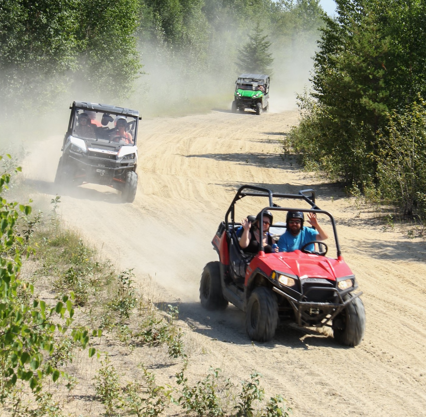 Side-by-sides with smiling riders come racing around a bend in a forest trail by Big Water Campground, kicking up dust behind them. 