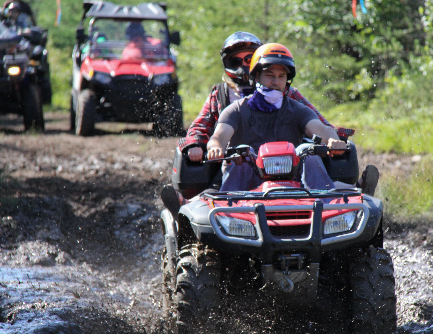 riders on ATVs splash through a mudhole in a forest trail on a sunny summer day at Big Water Campground.