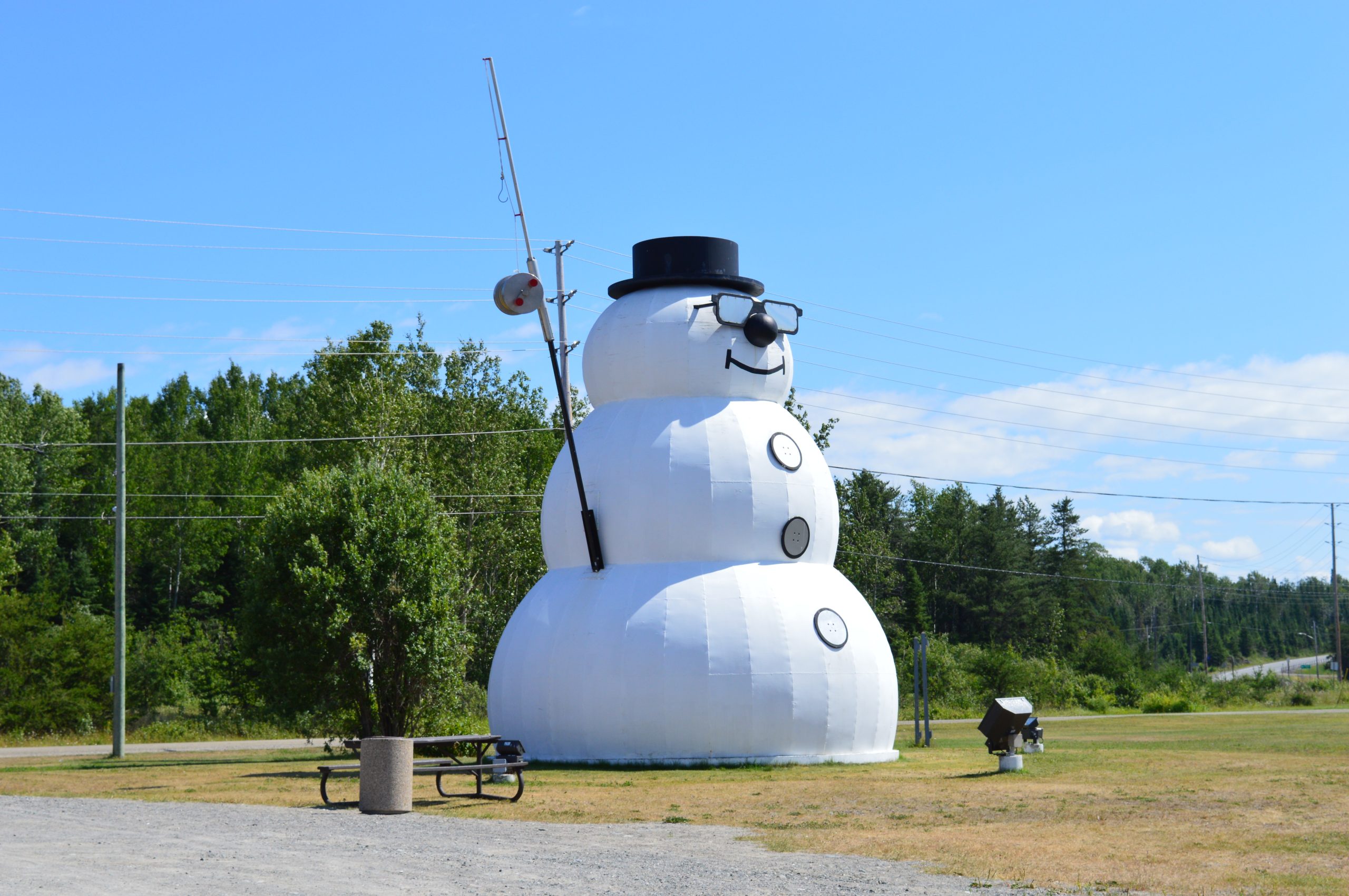the large metal statue, the Beardmore Snowman, sitting in a grassy field in front of a green forest. The snowman wears glasses, a pleasant smile, a black top hat, and holds a tall fishing pole.