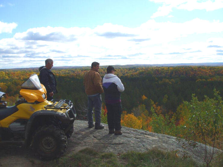 3 riders stand next to their ATV on a rock lookout by Booth Landing, gazing out at a wide forested valley and open sky. 