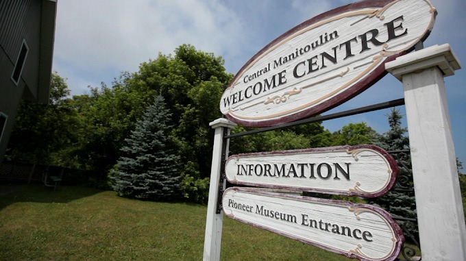 a carved wooden post sign in front of green trees, which reads "Central Manitoulin Welcome Centre", "Information", "Pioneer Museum Entrance".