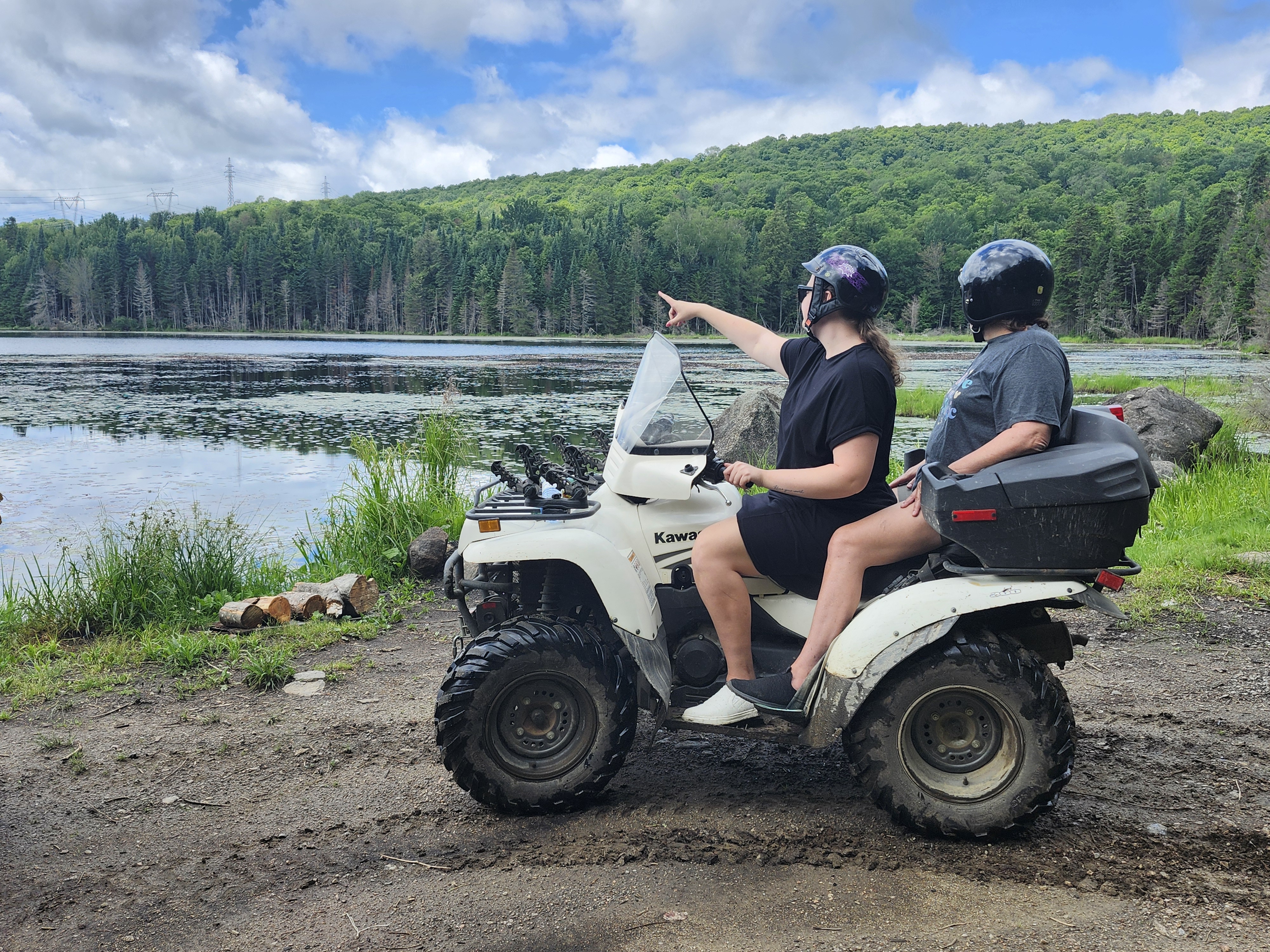 Two riders on an ATV sit beside a lake in the summer, looking to the distance at the forested opposite shore while one rider points. 