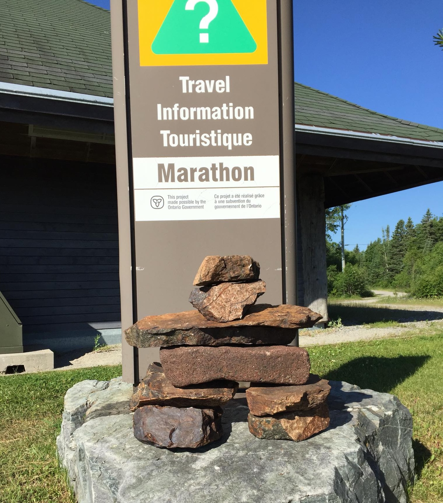 a little rock inukshuk sits in front of a brown pillar sign that reads "Travel Information Touristique Marathon".  A brown building, green grass and vivid blue sky are in the background.