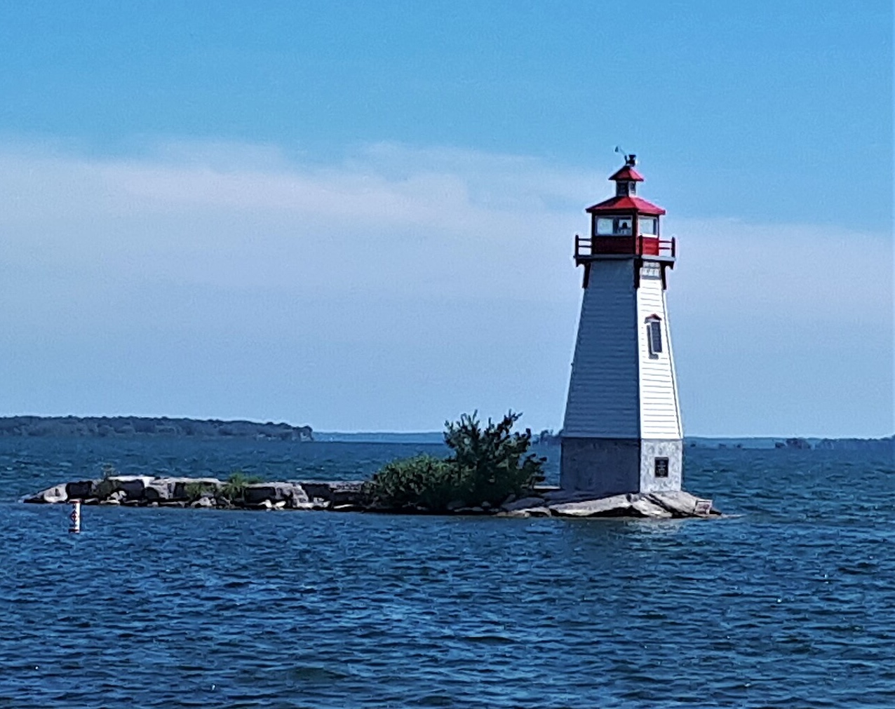 The red and white Jackson's Point lighthouse, sitting on a rocky shoal in the cool blue water of Lake Simcoe under a bright blue summer sky.