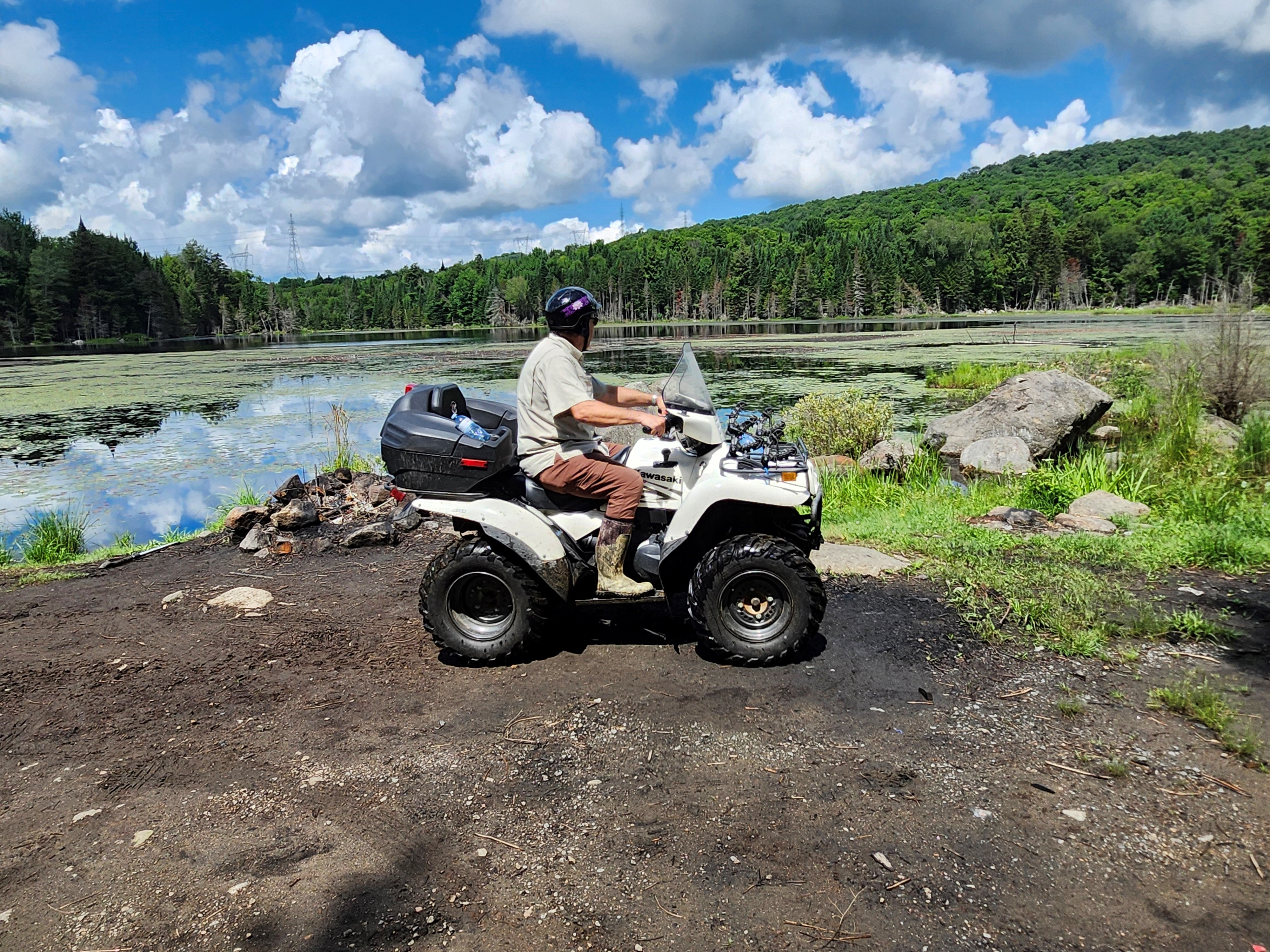 an ATV rider parked near a quiet lake in the summer, looking out over the glassy water and green forest. 