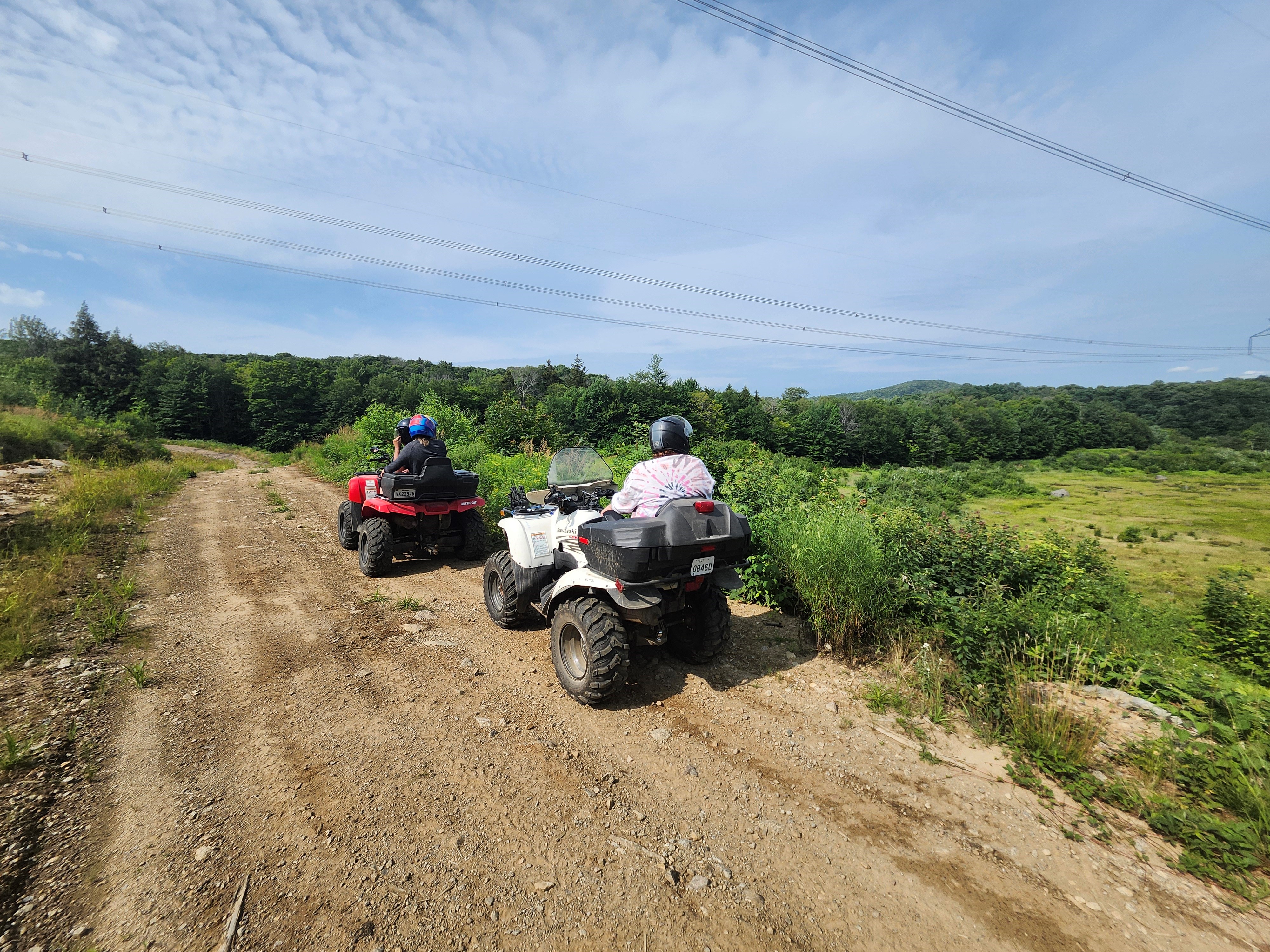 Two ATV riders drive their machines along a gravel trail through green fields dotted with trees under a soft blue summer sky.