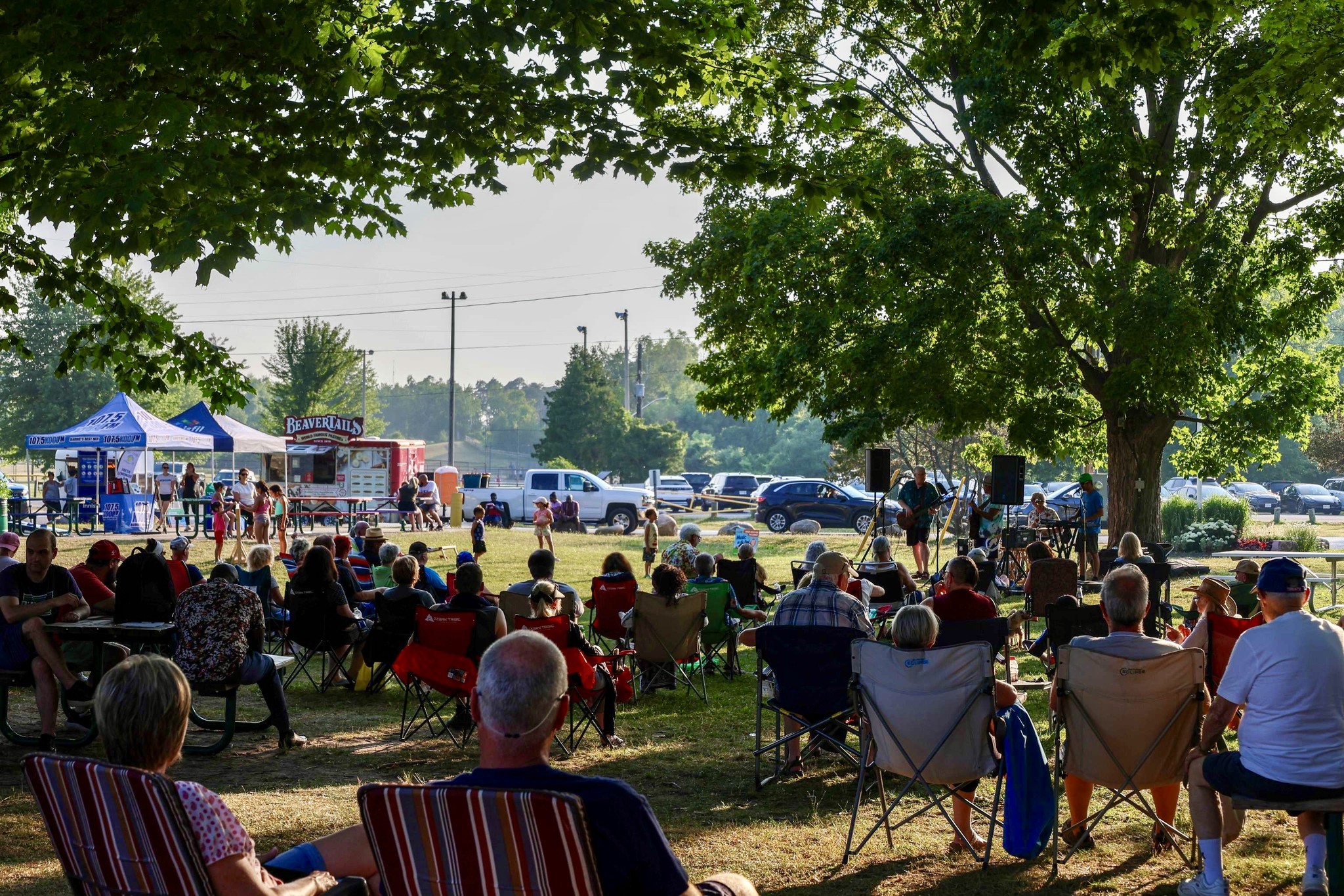 a crowd of people in lawn chairs sit under large shade trees watching live music on a summer day at Innisfil Beach Park. Food trucks and lakefront are in the background.