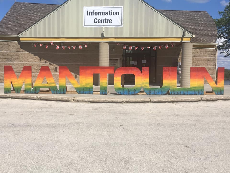 a wooden building with a peaked overhang which reads "information centre",  and a bright, rainbow-coloured "Manitoulin" sign in front made of large free-standing wooden letters.