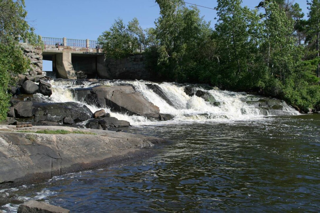 Nestor Falls, a low gentle falls passing under a bridge near the town, surrounded by green forest. It bubbles into a clear calm riverpool with smooth rock shores. 