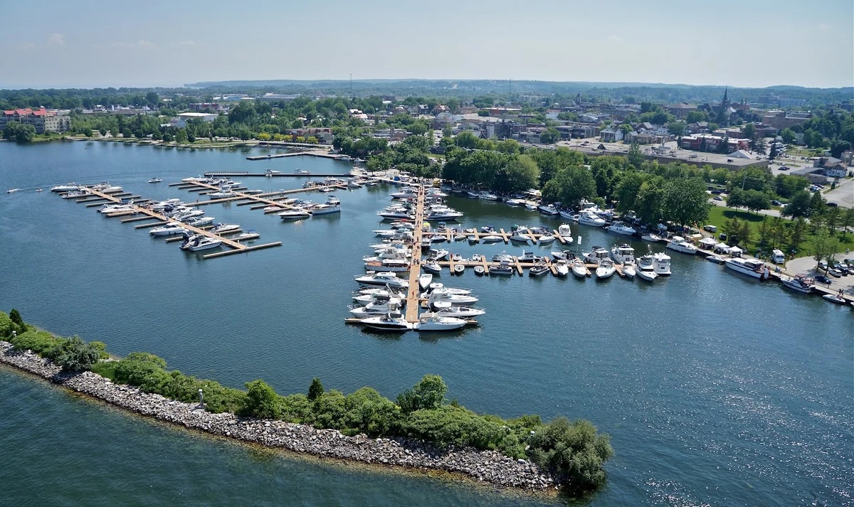 boats fill the many slips on the calm blue water at Port of Orillia. Lush green trees dot the bustling lakeshores and the summer sky is clear and blue.