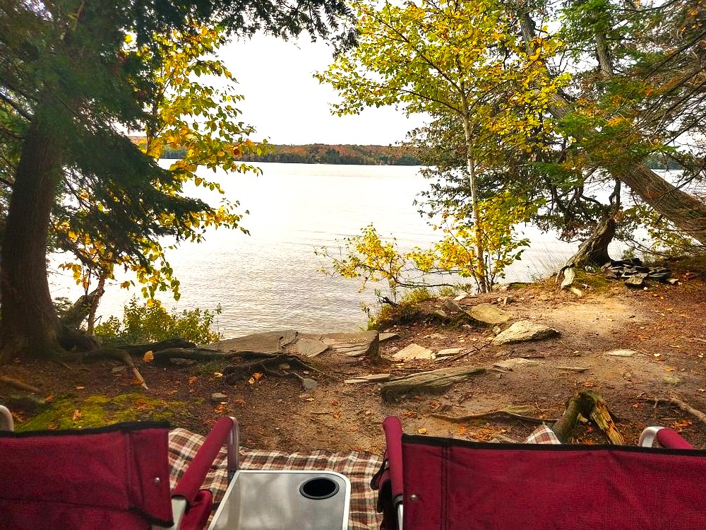 folding camp chairs and a table in a shady campsite facing out toward a quiet lake at Restoule Provincial Park. 
