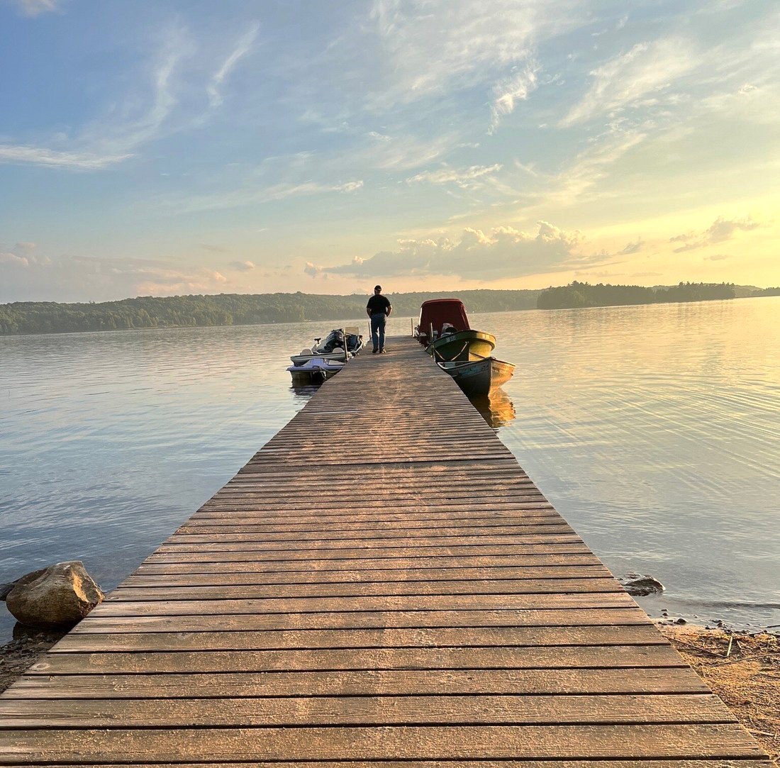 a long wooden dock leading out into a calm, shining lake at sunrise at Restoule Provincial Park. A person walks to a boat at the end of the dock. 