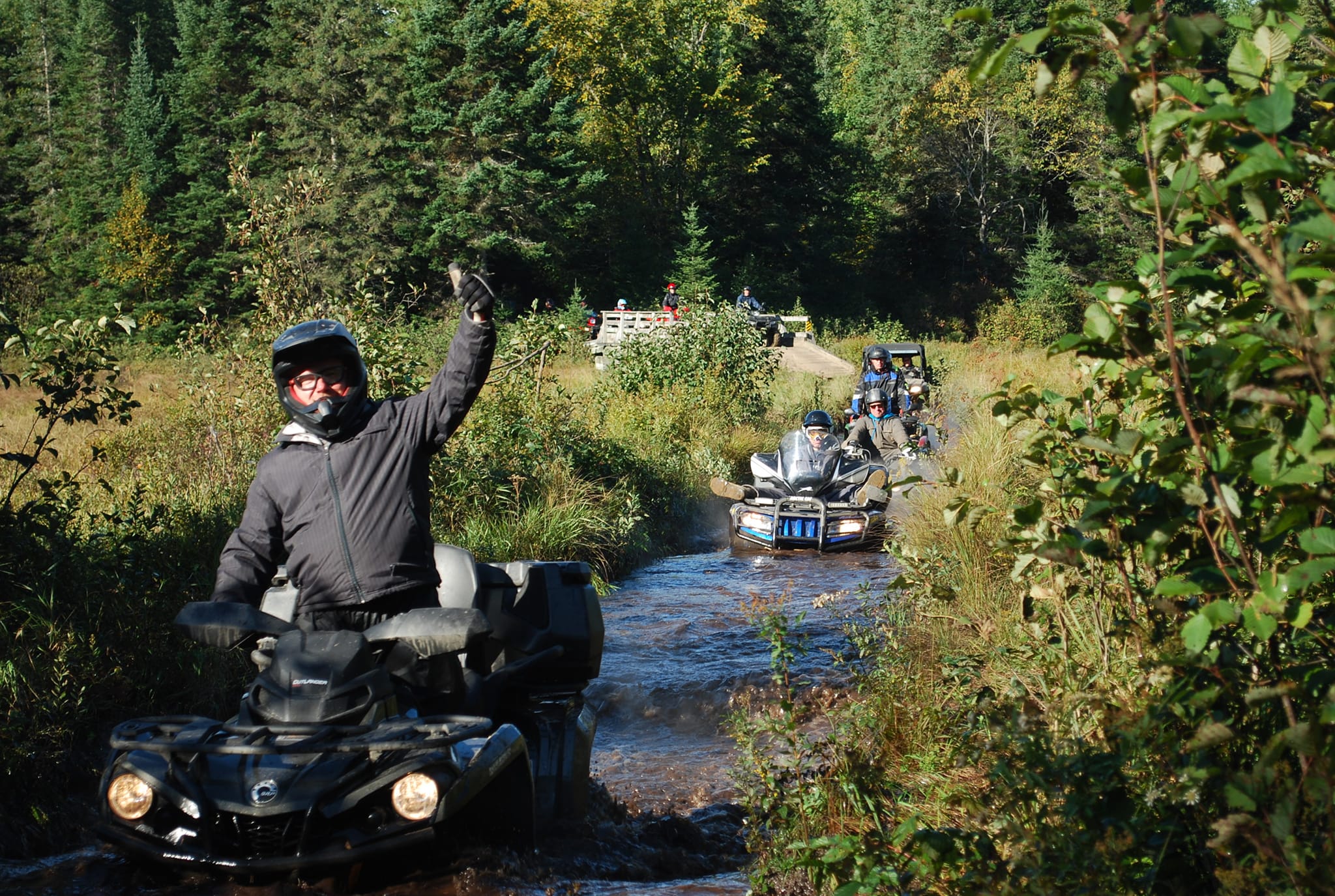 a line of ATV riders drive through a flooded creek bed in a clearing in the forest. The rider at the front gives a smiling thumbs up as they ride through water up to their headlights.