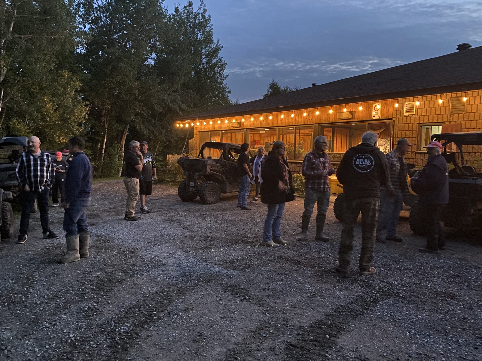 The Elliot Lake ATV Club members happily mingle in the parking lot of the clubhouse under an illuminated awning on a summer evening.