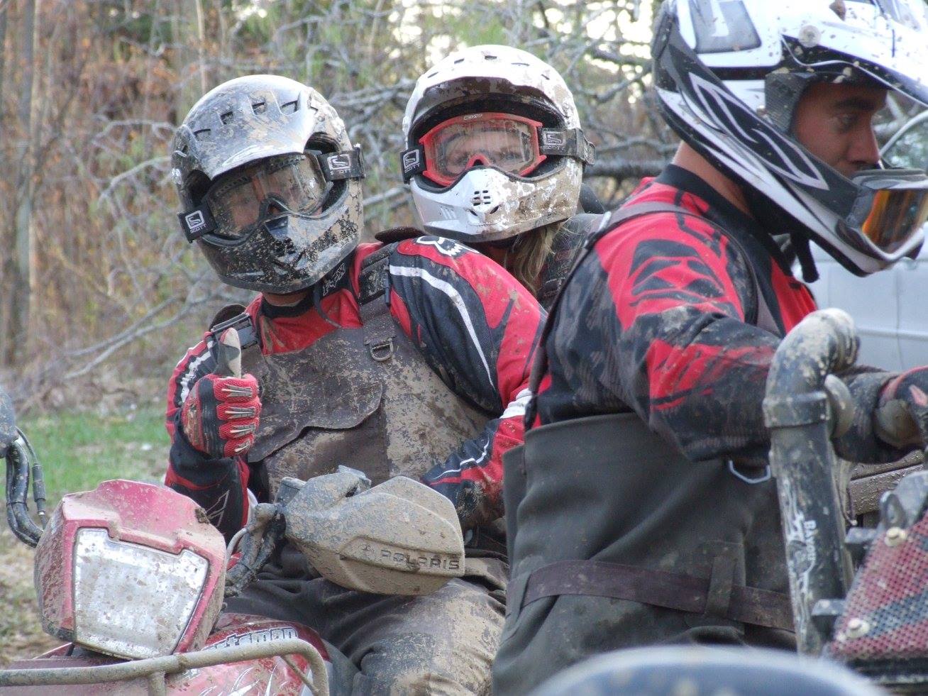 muddy ATV riders in helmets on a forest trail. One rider flashes a thumbs up. 