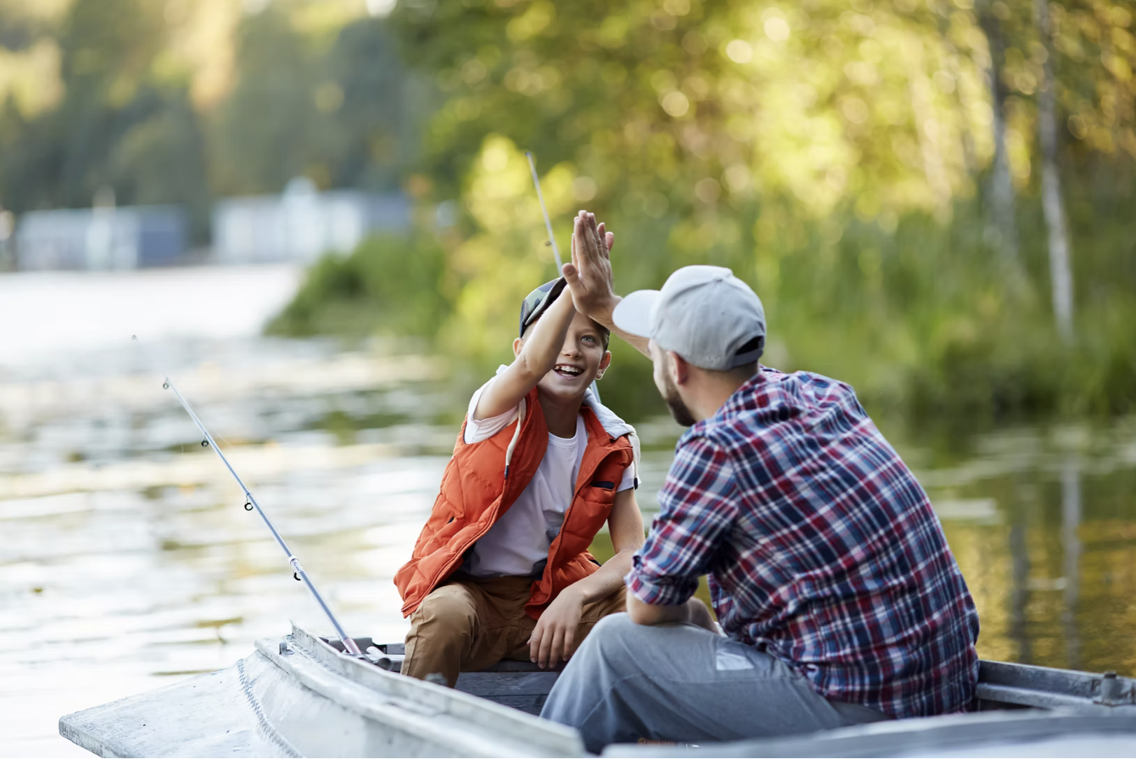 a father and son smile and high-five as they sit in a rowboat with their fishing rods on a summer day. 