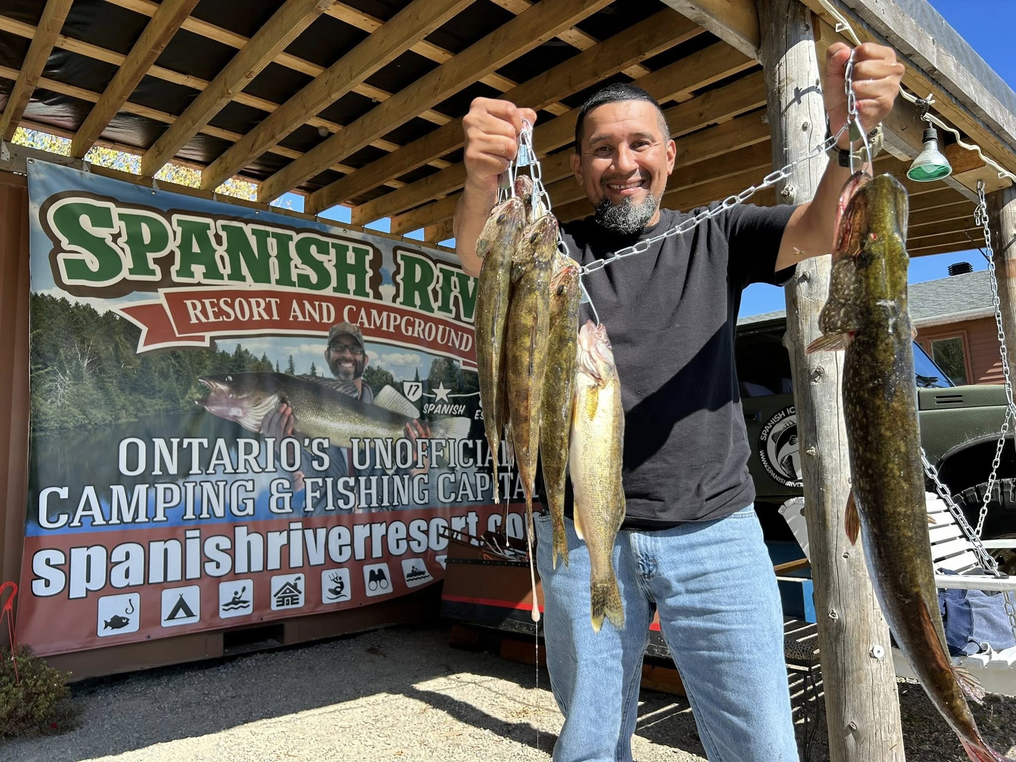 a smiling angler holds up a chain of several large fish next to a sign that reads "Spanish River Resort and Campground".