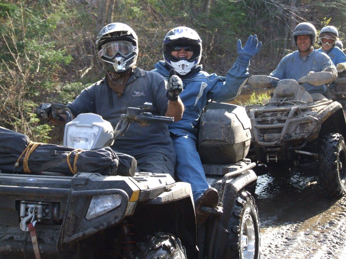 A line of riders on ATVs drive down a muddy forest trail. One rider waves happily. 
