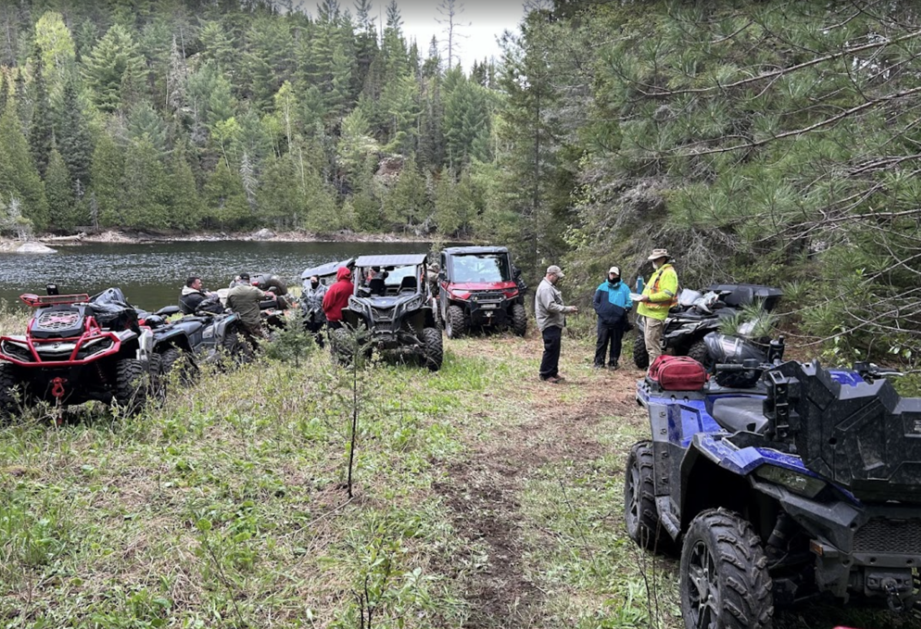 The West Nipissing ATV Club members chat on a thickly forested riverbank next to their parked ATVs.