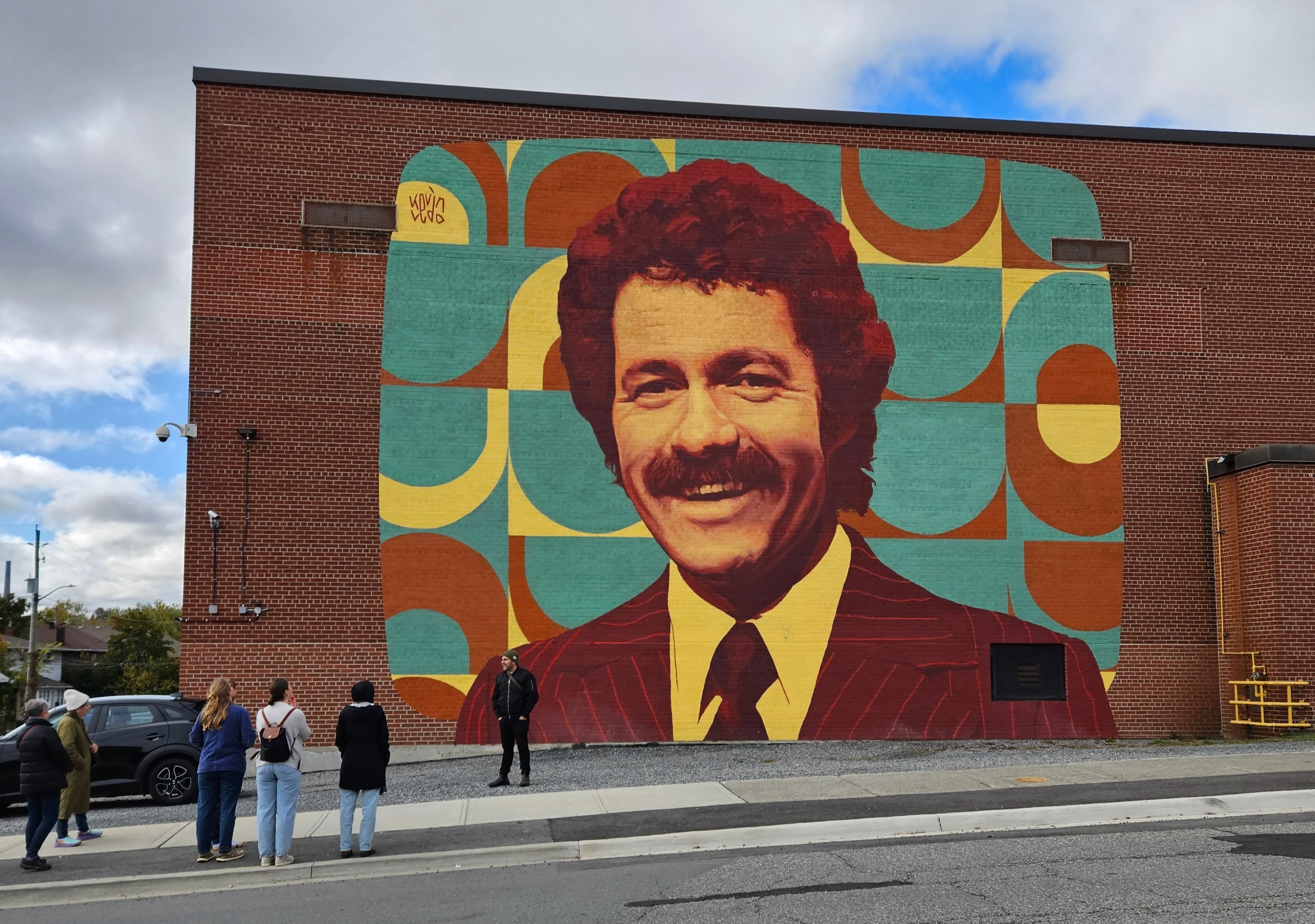 A turquoise and brown vintage-seventies-inspired mural of Alex Trebek in his younger days. It covers the full side of a brick high school in Sudbury, and visitors on a tour stand to admire it.