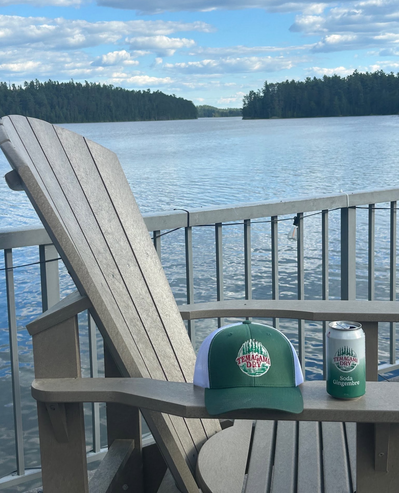A can of Temagami Dry ginger ale and an official hat bearing the Temagami Dry logo sit on the wooden arm of an adirondak chair on a deck overlooking a calm blue lake. 
