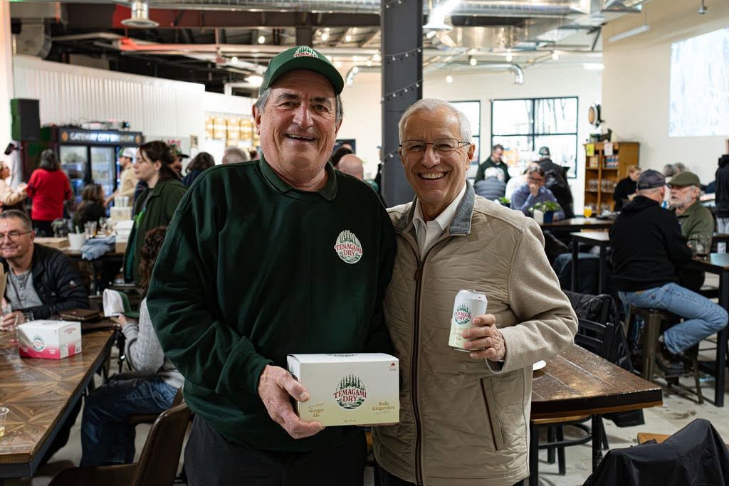 Mayor of Temagami Dan O'Mara smiles as he holds up a case of Temagami Dry ginger ale in a crowded brewery.