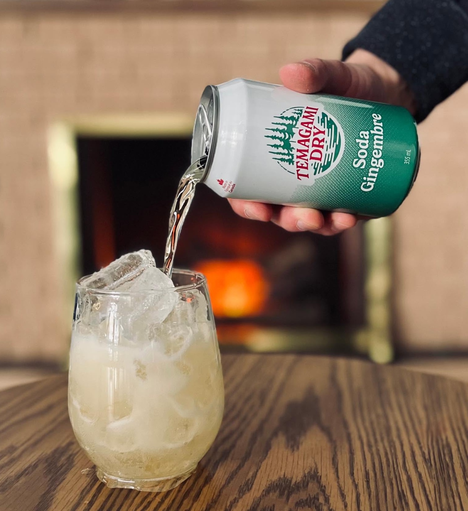a hand pours a can of Temagami Dry ginger ale into a glass full of ice on a wooden table in front of a glowing indoor fireplace.