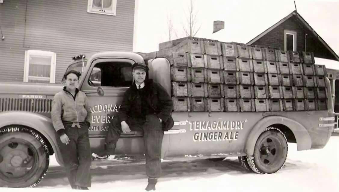 a black and white vintage photograph of two men posing in front of a pickup truck bearing the "Temagami Dry" label and loaded with cases of the iconic soda.