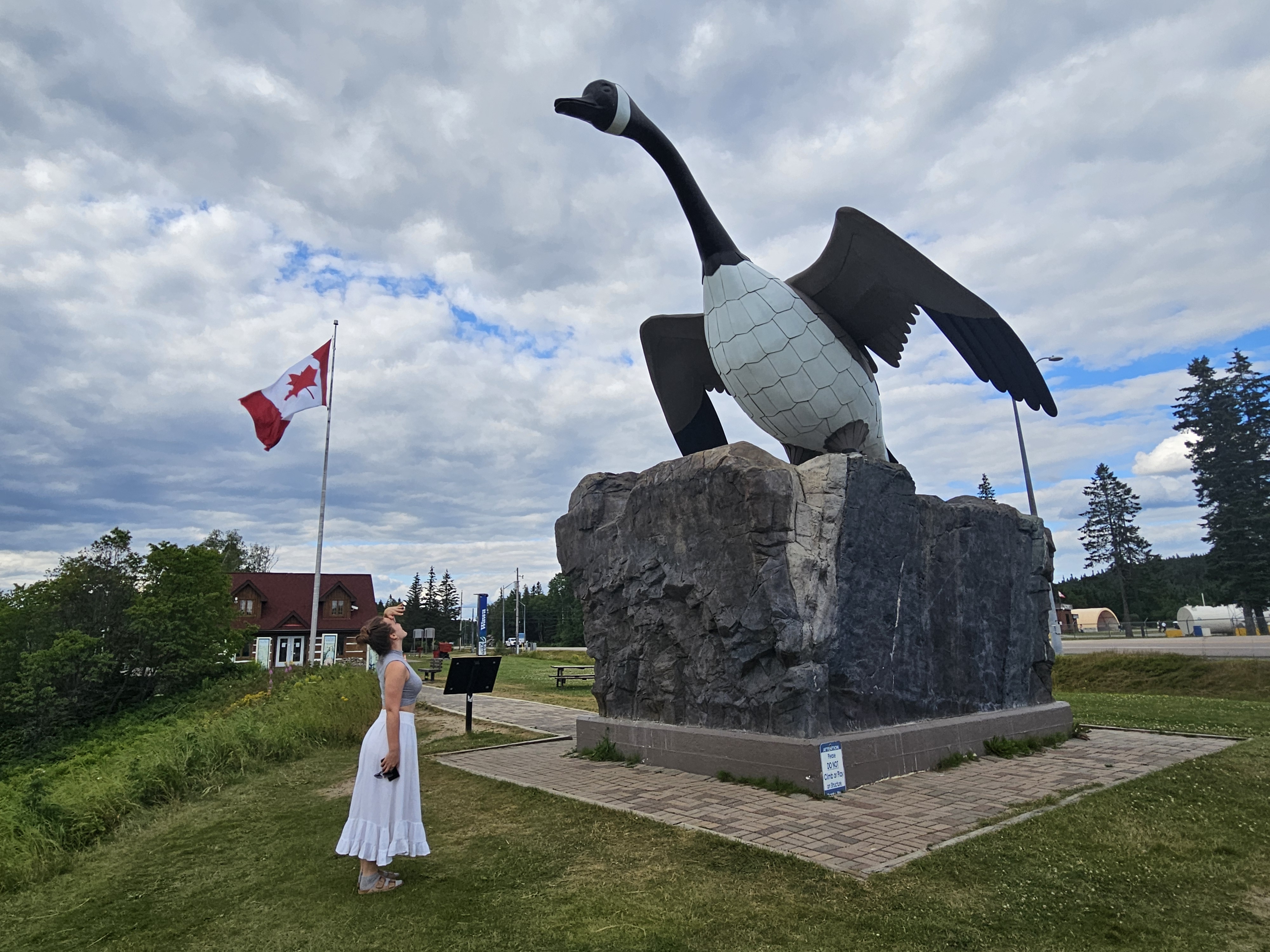 a smiling woman in a long white skirt holds her hand like a visor above her eyes and looks way up at the Wawa Goose statue towering above her. Green grass, trees and a few buildings sit in the background.