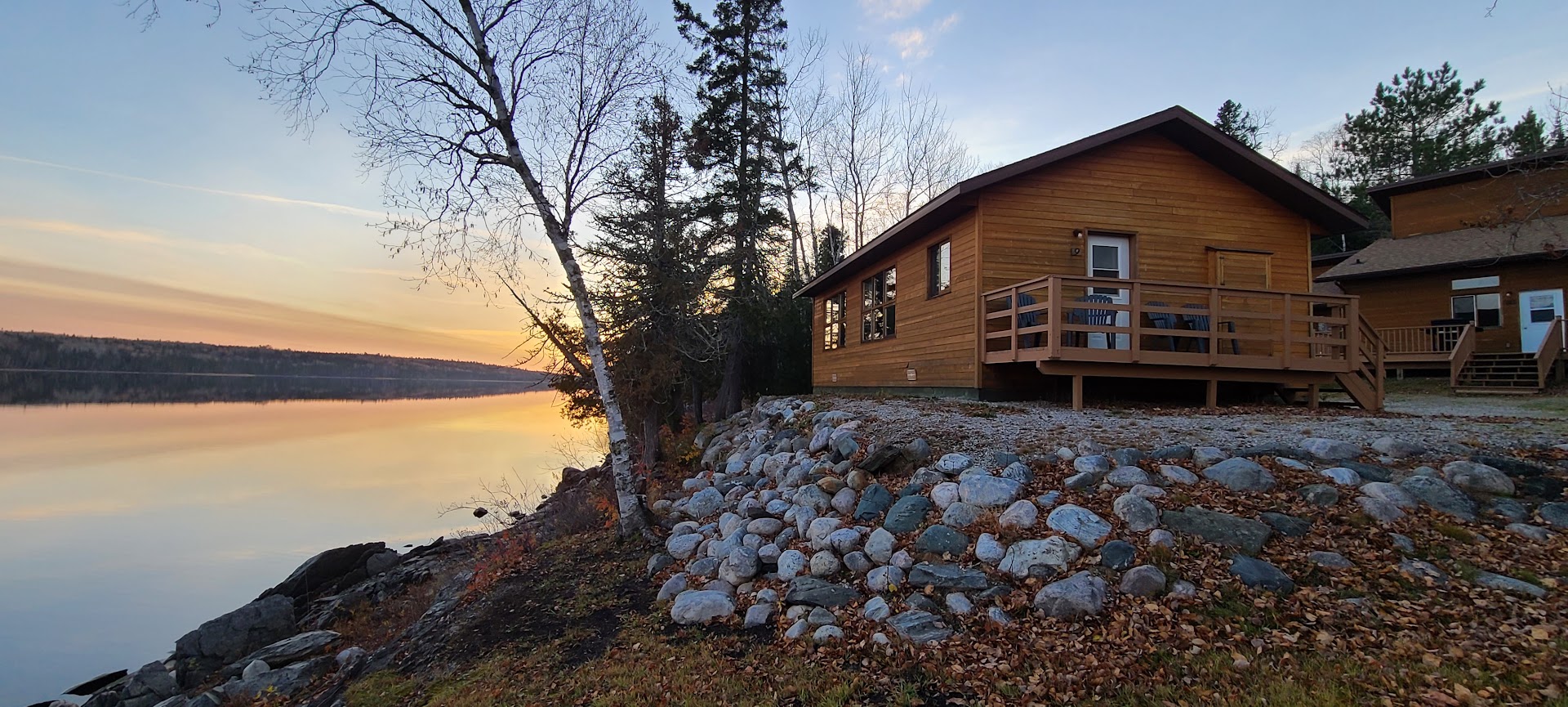 A wooden cabin at Anderson's Lodge on a rocky, forested shore next to a calm, glassy lake lit up with an orange sunset. 