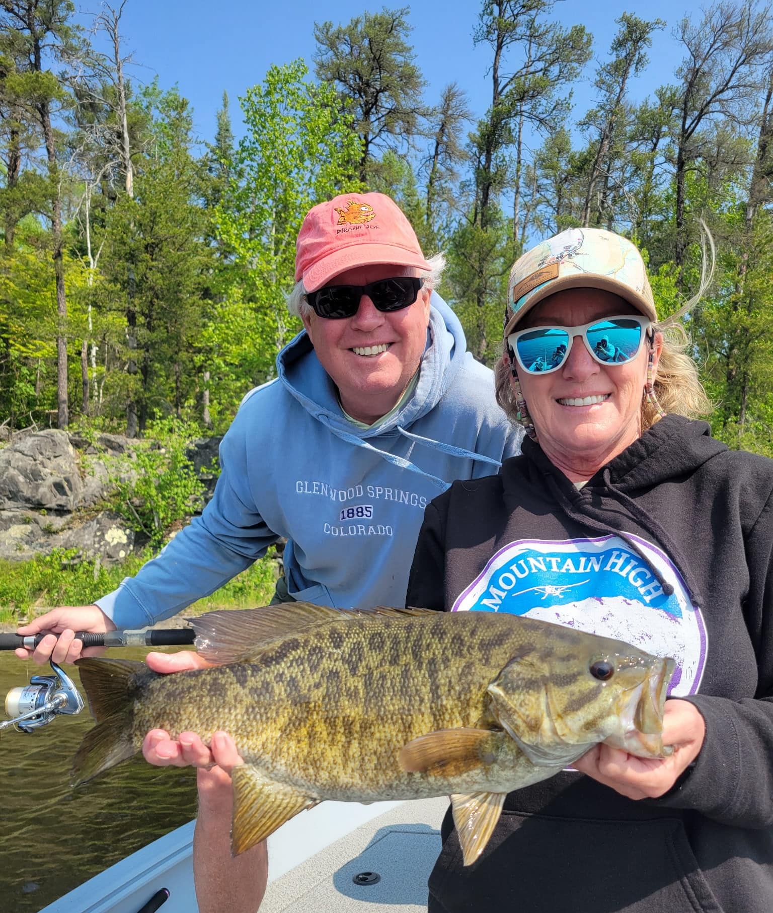 a smiling, older man and woman wearing hats and sunglasses stand in their fishing boat holding up a large fish. 